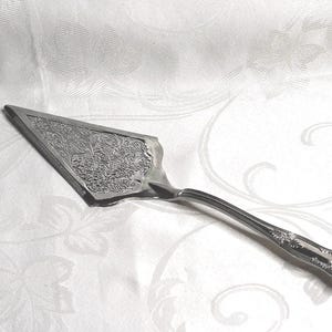May include: A silver cake server with an ornate, floral design on the blade and handle. The triangular blade is detailed with a repeating pattern. The handle curves gently and has a decorative end. The server rests on a white, patterned tablecloth.