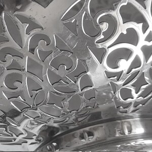 May include: A polished silver-coloured metal bowl featuring an intricate, openwork filigree pattern with swirling scroll motifs and geometric cutouts. The bowl has a smooth, reflective surface and a circular base, showcasing a detailed decorative design.