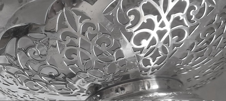 May include: A polished silver-coloured metal bowl featuring an intricate, openwork filigree pattern with swirling scroll motifs and geometric cutouts. The bowl has a smooth, reflective surface and a circular base, showcasing a detailed decorative design.