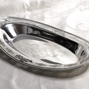 May include: An oval-shaped silver-coloured metal dish with a polished, reflective surface. The wide rim features an engraved decorative pattern consisting of repeating vertical lines and floral motifs spaced evenly around the entire perimeter.