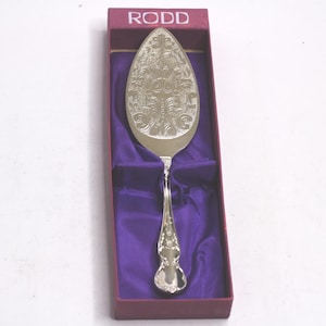May include: Silver cake server with an engraved pattern, displayed in a burgundy box with a purple satin lining. The box is branded with the name "RODD" in white lettering.