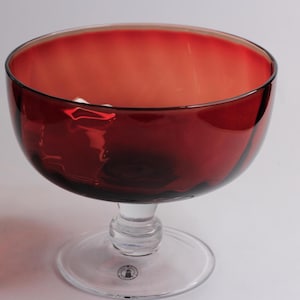 May include: A deep red glass bowl with a clear glass pedestal base. The bowl has a wide, rounded form and a smooth, glossy finish. The base is circular and provides stability. This elegant piece is suitable for serving or display.