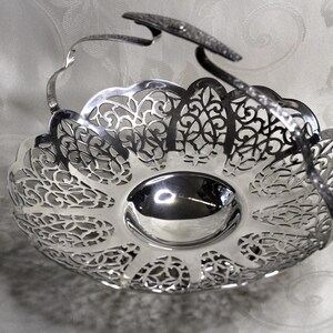 May include: A silver-coloured metal basket featuring a polished, smooth circular base and an intricate, openwork filigree rim with a scalloped edge. The basket includes a single, arched, textured handle that extends across the top.