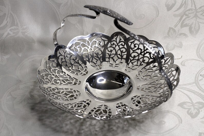 May include: A silver-coloured metal basket featuring a polished, smooth circular base and an intricate, openwork filigree rim with a scalloped edge. The basket includes a single, arched, textured handle that extends across the top.