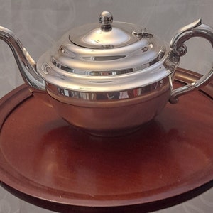 May include: A silver teapot with a curved spout and handle, on a round, dark brown wooden tray. The teapot has a lid with a small knob. The handle has a decorative, curved design. The tray is a deep reddish-brown colour.