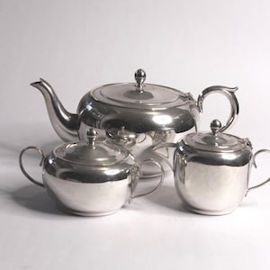 May include: A silver-toned tea set consisting of a large teapot with a curved spout and handle, a lidded sugar bowl with two side handles, and a lidded creamer with a single handle. Each piece features a smooth, reflective metallic finish.