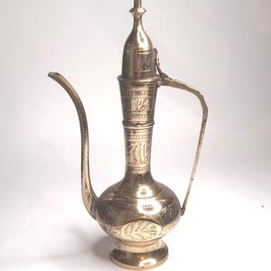 May include: A vintage brass pitcher with an elongated spout and handle. The pitcher features intricate etched floral and geometric patterns. The design includes a bulbous base and a tall, slender neck with a lid. The overall colour is a warm, antique gold.