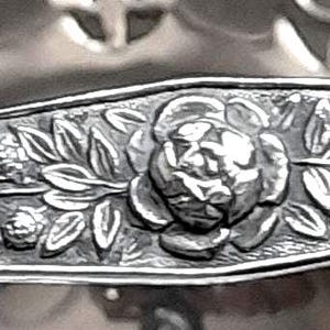 May include: A silver-toned metal handle featuring an embossed floral pattern with a central rose motif surrounded by leaves and small buds. The handle has a tapered shape and is positioned over a decorative bowl with a pierced lattice design.