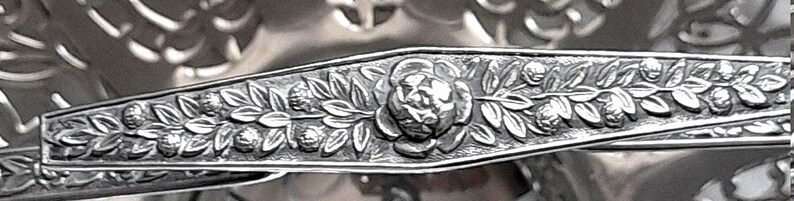 May include: A silver-toned metal handle featuring an embossed floral pattern with a central rose motif surrounded by leaves and small buds. The handle has a tapered shape and is positioned over a decorative bowl with a pierced lattice design.