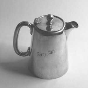 May include: A silver coffee pot with a curved handle and a spout. The lid has a small knob on top. The words "Savoy Cafe" are engraved on the side. The pot is likely made of metal, suitable for serving coffee.