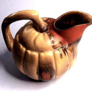 May include: A ceramic jug with a pumpkin-like design. The jug is mainly tan with brown and reddish-brown accents. It features a handle and a spout, and the overall shape is rounded.