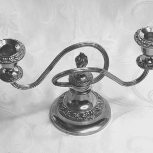 May include: A silver candelabra with two candle holders. The candelabra has a decorative base and a curved arm connecting the candle holders. The candle holders have ornate detailing around the rims. The candelabra is made of metal.