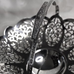 May include: A silver-toned metal basket featuring an intricate openwork filigree pattern along the scalloped rim and a central solid circular base. The basket includes a single upright handle decorated with a repeating floral vine engraving.