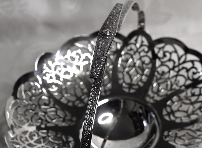 May include: A silver-toned metal basket featuring an intricate openwork filigree pattern along the scalloped rim and a central solid circular base. The basket includes a single upright handle decorated with a repeating floral vine engraving.