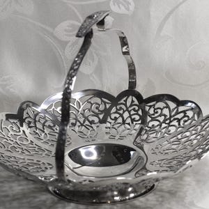 May include: A polished silver-tone metal basket featuring an intricate openwork filigree pattern along the scalloped rim and a single arched handle. The base is circular and solid, supporting the wide, shallow bowl with its detailed cut-out design.