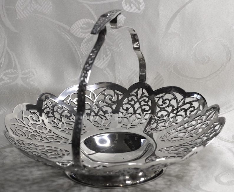 May include: A polished silver-tone metal basket featuring an intricate openwork filigree pattern along the scalloped rim and a single arched handle. The base is circular and solid, supporting the wide, shallow bowl with its detailed cut-out design.