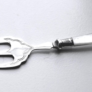 May include: A silver serving fork with three prongs and a decorative handle. The handle has a white finish and a small emblem. The fork is likely for serving appetizers or desserts. The handle is possibly mother-of-pearl.