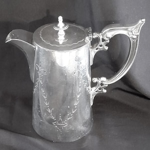May include: A silver-toned metal teapot featuring a hinged lid with a small finial, a curved spout, and an ornate, scrolled handle. The cylindrical body is decorated with delicate, etched floral and vine patterns on the front surface.