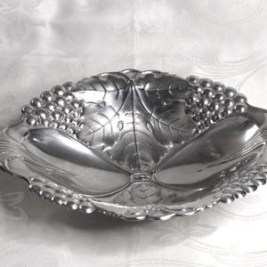 May include: A polished silver-toned metal bowl featuring a decorative embossed pattern of grape clusters and large veined leaves around the rim. The interior of the bowl has a smooth, reflective surface with a scalloped edge and a circular base.