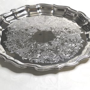 May include: A silver-coloured, oval serving tray with an ornate floral pattern. The tray has a scalloped edge and a reflective surface, suitable for serving food or displaying items. The tray is made of metal.