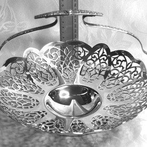 May include: A silver-toned metal basket with a polished finish, featuring an intricate openwork filigree pattern along the sides and a swing handle. A vertical metal ruler is positioned behind the basket, showing a height of approximately 25 cm.