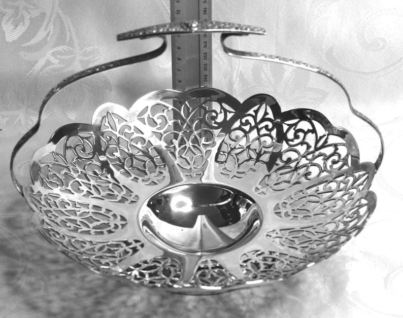 May include: A silver-toned metal basket with a polished finish, featuring an intricate openwork filigree pattern along the sides and a swing handle. A vertical metal ruler is positioned behind the basket, showing a height of approximately 25 cm.