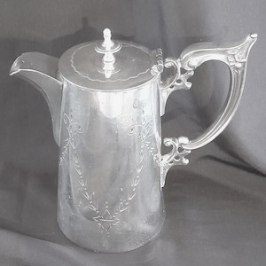 May include: A silver-toned metal coffee pot featuring a cylindrical body with an engraved floral garland pattern, a hinged lid with a small finial, a curved spout, and an ornate, scrolled handle. The surface has a polished, reflective finish.