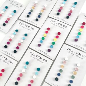 May include: Assortment of colorful small round earrings displayed on white cards with the text "THE YUM CO. Jewellery & Accessories" and the company website address.