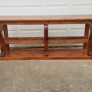 Op de afbeelding: Een lange, houten consoletafel met twee planken en decoratieve krulwerk op de poten. De tafel is in een rijke bruine kleur gebeitst.