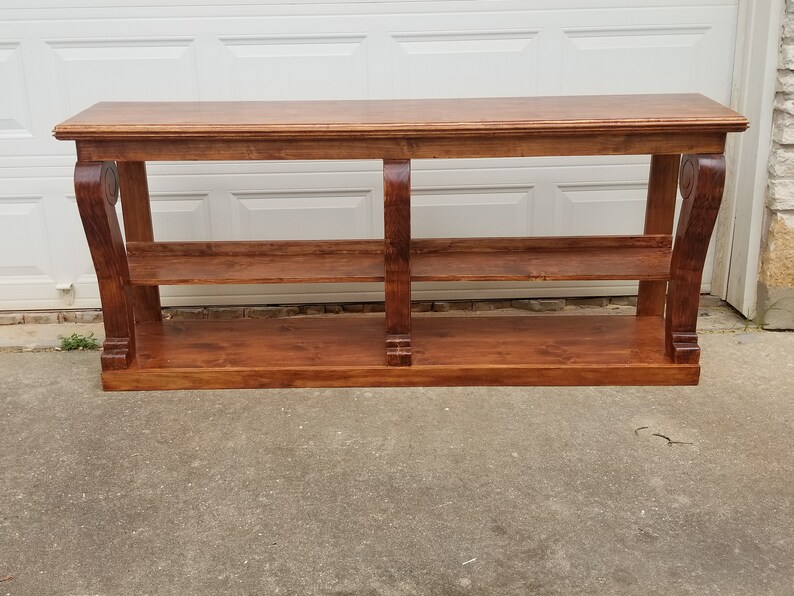Op de afbeelding: Een lange, houten consoletafel met twee planken en decoratieve krulwerk op de poten. De tafel is in een rijke bruine kleur gebeitst.