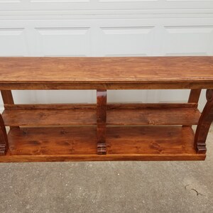 Op de afbeelding: Een houten consoletafel met twee planken en decoratieve, gerolde poten. De tafel is in een rijke bruine kleur gebeitst.