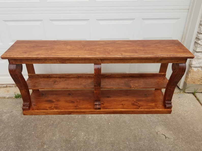 Op de afbeelding: Een houten consoletafel met twee planken en decoratieve, gerolde poten. De tafel is in een rijke bruine kleur gebeitst.
