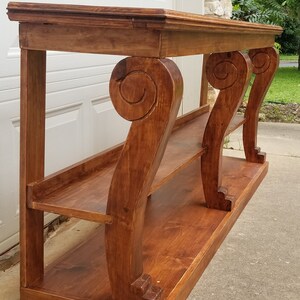 Può includere: Un tavolo console in legno con tre gambe ornate e due ripiani. Il tavolo è tinto di un colore marrone intenso.