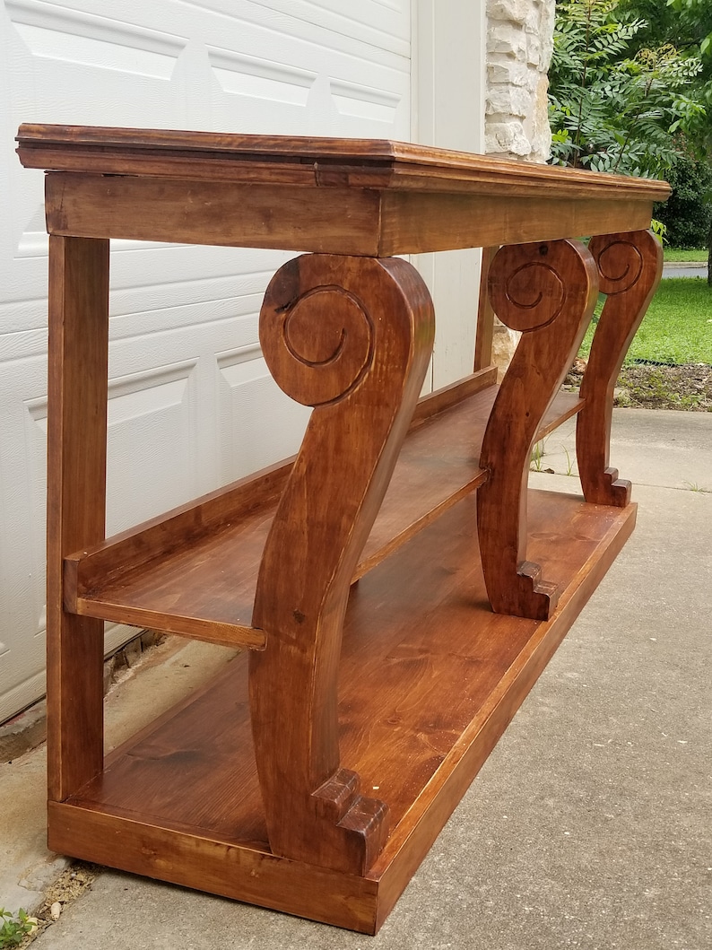 Op de afbeelding: Een houten consoletafel met drie sierlijke poten en twee planken. De tafel is in een rijke bruine kleur gebeitst.