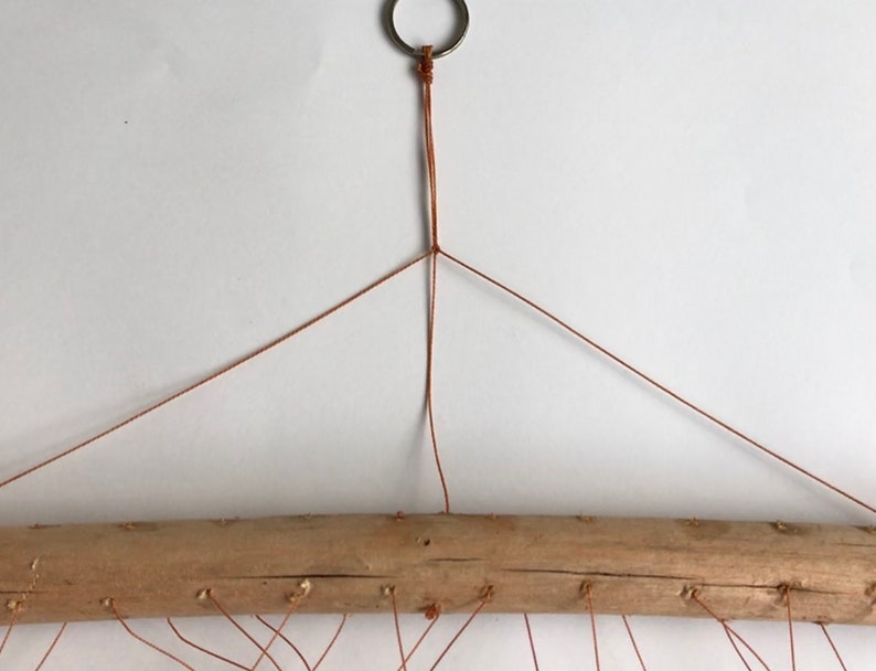 May include: A brown cord is tied to a metal ring and then to a wooden branch. The cord is strung across the branch to create a hanging macrame design.