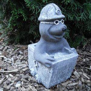 Könnte beinhalten: Eine graue Gartenfigur aus Beton, die eine Maulwurf darstellt, der einen Bergmannhelm und eine Schutzbrille trägt und in einem kleinen grauen Beton-Bergwerkswagen sitzt.