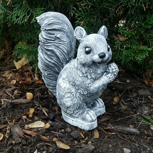 Könnte beinhalten: Eine graue Beton-Eichhörnchen-Statue mit buschigem Schwanz, die auf einem Bett aus braunem Mulch sitzt. Das Eichhörnchen hält eine kleine Eichel in seiner Pfote.