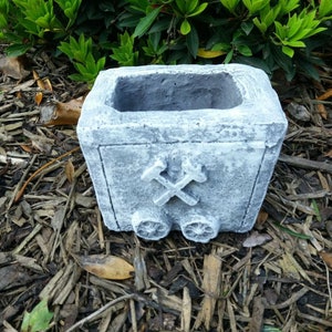 Könnte beinhalten: Ein grauer Beton-Pflanztopf in Form eines Grubenwagens mit einem Design aus Spitzhacke und Schaufel. Der Topf steht auf einem Bett aus braunem Mulch.