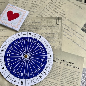 May include: A blue and white alphabet wheel with a gold pointer. The wheel is surrounded by old, faded paper documents with handwritten text.
