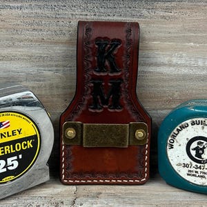 May include: A brown leather tape measure holder with the initials "KM" embossed on the front. A Stanley Powerlock 25' tape measure and a teal tape measure are on either side of the holder.