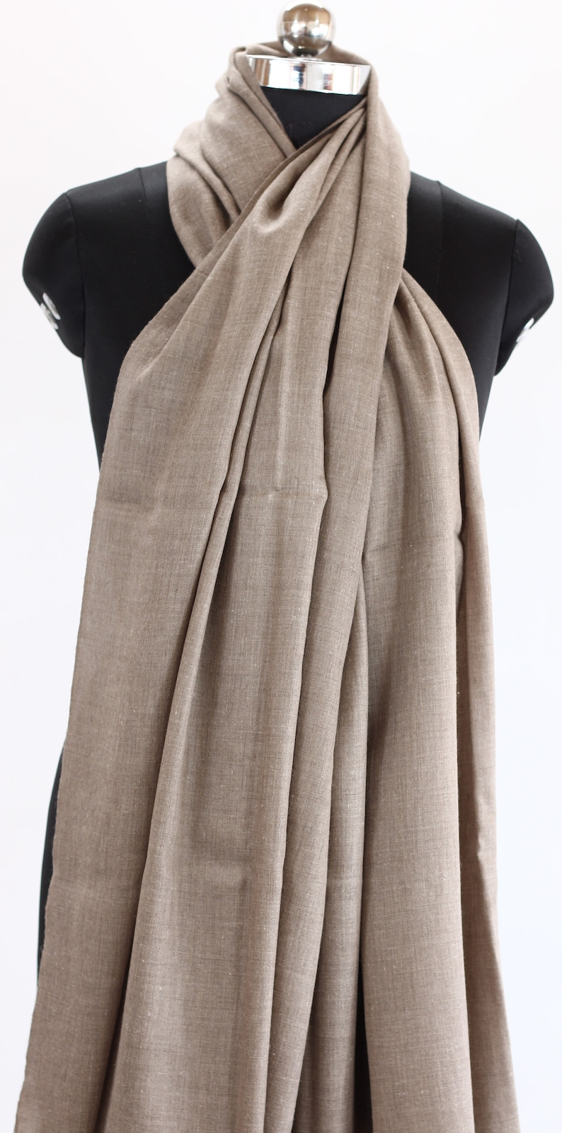 May include: A light brown pashmina scarf draped over a black mannequin. The scarf is made of a soft, woven fabric and has a subtle sheen.