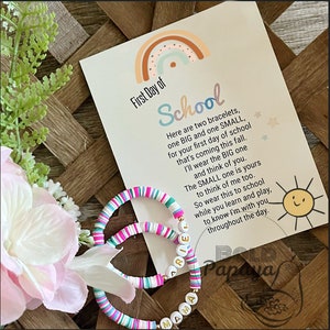 Puede incluir: Dos pulseras de cuentas coloridas con las palabras "Mama" y "Ariel" escritas en letras doradas. Las pulseras están sobre una tarjeta blanca con un arcoíris y el texto "Primer día de escuela" y un poema sobre las pulseras.