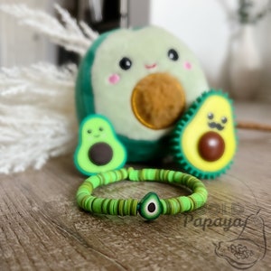 Puede incluir: Pulsera verde en forma de aguacate con cuentas verdes y marrones. La pulsera tiene un pequeño dije de aguacate con una semilla marrón en el centro. La pulsera está rodeada de tres juguetes de peluche en forma de aguacate.