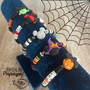 May include: Five Halloween-themed bracelets with different character charms, including a witch, a ghost, and a pumpkin. The bracelets are displayed on a blue velvet stand. The background is a white spiderweb.
