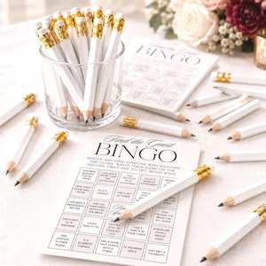 May include: A clear glass cup filled with white pencils featuring gold ferrules and erasers, alongside printed Find the Guest BINGO game cards. The cards contain a grid of squares with text prompts for guests to mingle and find people who match clues.