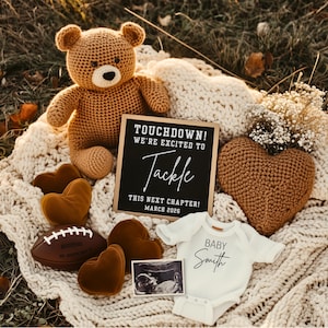 Op de afbeelding: Een aankondigingsscène voor een zwangerschap met een bruine teddybeer, een voetbal en een bord met de tekst "TOUCHDOWN! WE'RE EXCITED TO TACKLE THIS NEXT CHAPTER! MAART 2026." Een babyromper en een echo completeren de scène.