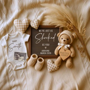 May include: A baby announcement featuring a white onesie with the name "Smith", a sonogram photo, and a letterboard with the text "We're just as shocked as you! Arriving March 2026". Includes a teddy bear, booties, and heart-shaped decor.