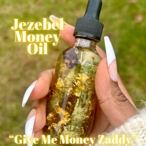 May include: A clear glass bottle labeled "Jezebel Money Oil" with a black dropper top. The bottle contains gold flakes and what appears to be a dollar bill. The text "Give Me Money Zaddy" is at the bottom of the bottle.