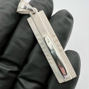May include: Silver pendant with a rectangular shape and the words "AITS Concrete" engraved on it.