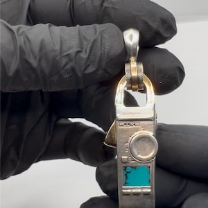 May include: Silver and gold pendant charm with a turquoise blue digital display and the word "FLUKE" printed on the front. The pendant has a round knob and a small silver triangle on the side.
